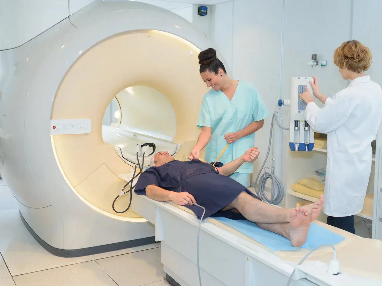 Pacjent leży w aparacie rezonansu magnetycznego, przygotowywany do badania. Personel medyczny monitoruje parametry, dbając o bezpieczeństwo i minimalizując ewentualne skutki uboczne rezonansu magnetycznego.