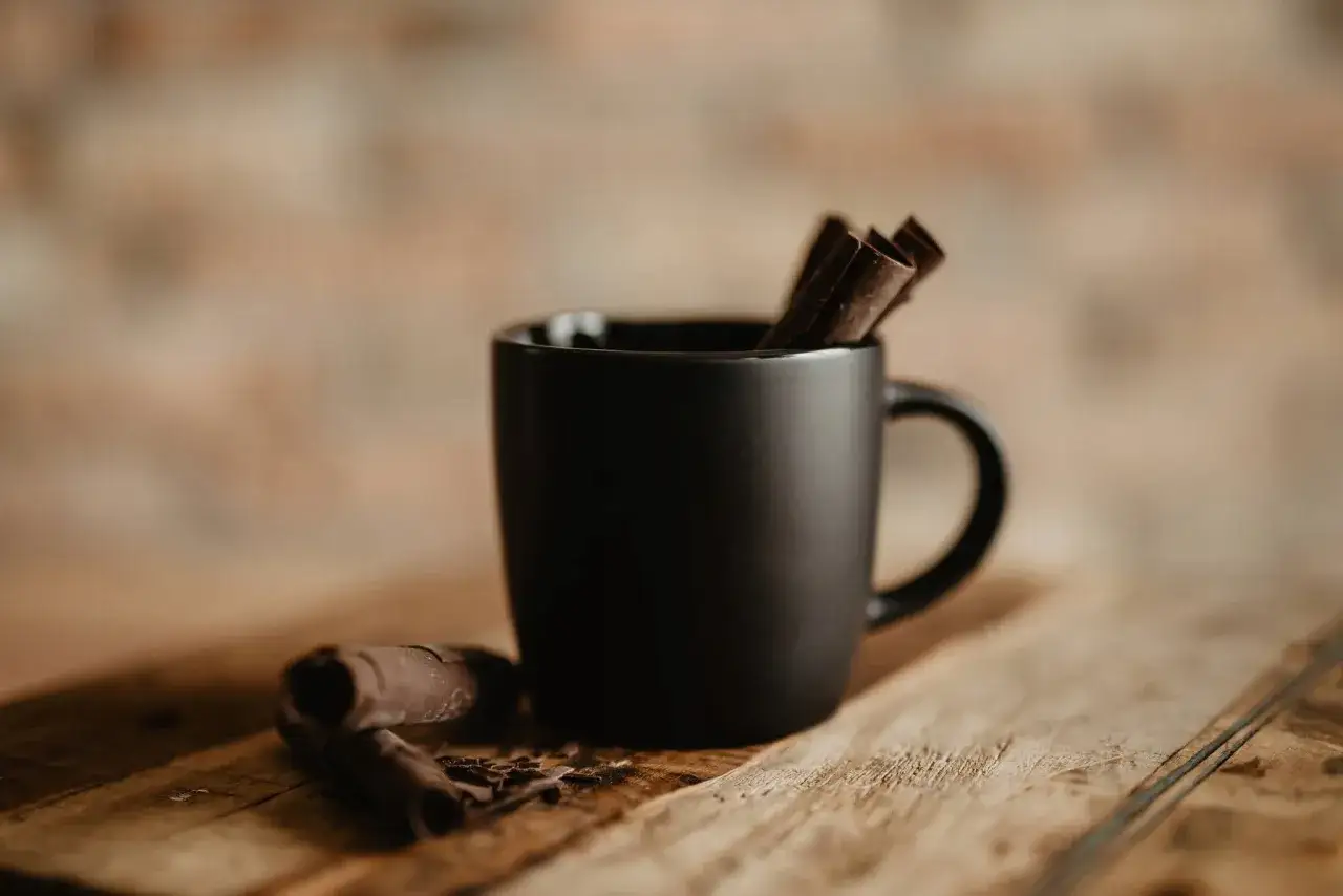 Czarny matowy kubek z pałeczkami cynamonu i czekoladowymi wiórkami. Trudno określić, jaką pojemność ma kubek, ale wygląda na idealny na gorącą czekoladę.