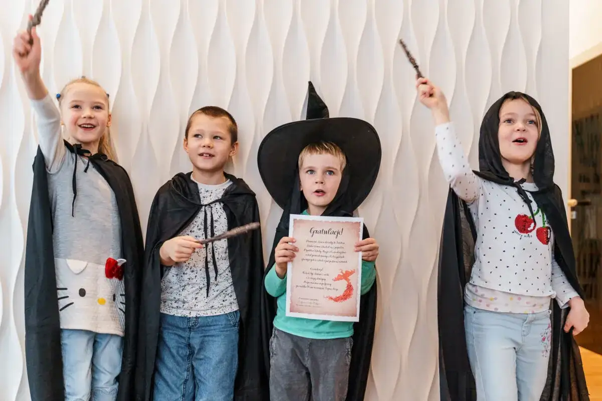 Dzieci w strojach czarodziejów, z różdżkami i pelerynami, pokazują, jak zrobić przebranie na Halloween. Jeden z chłopców trzyma dyplom.
