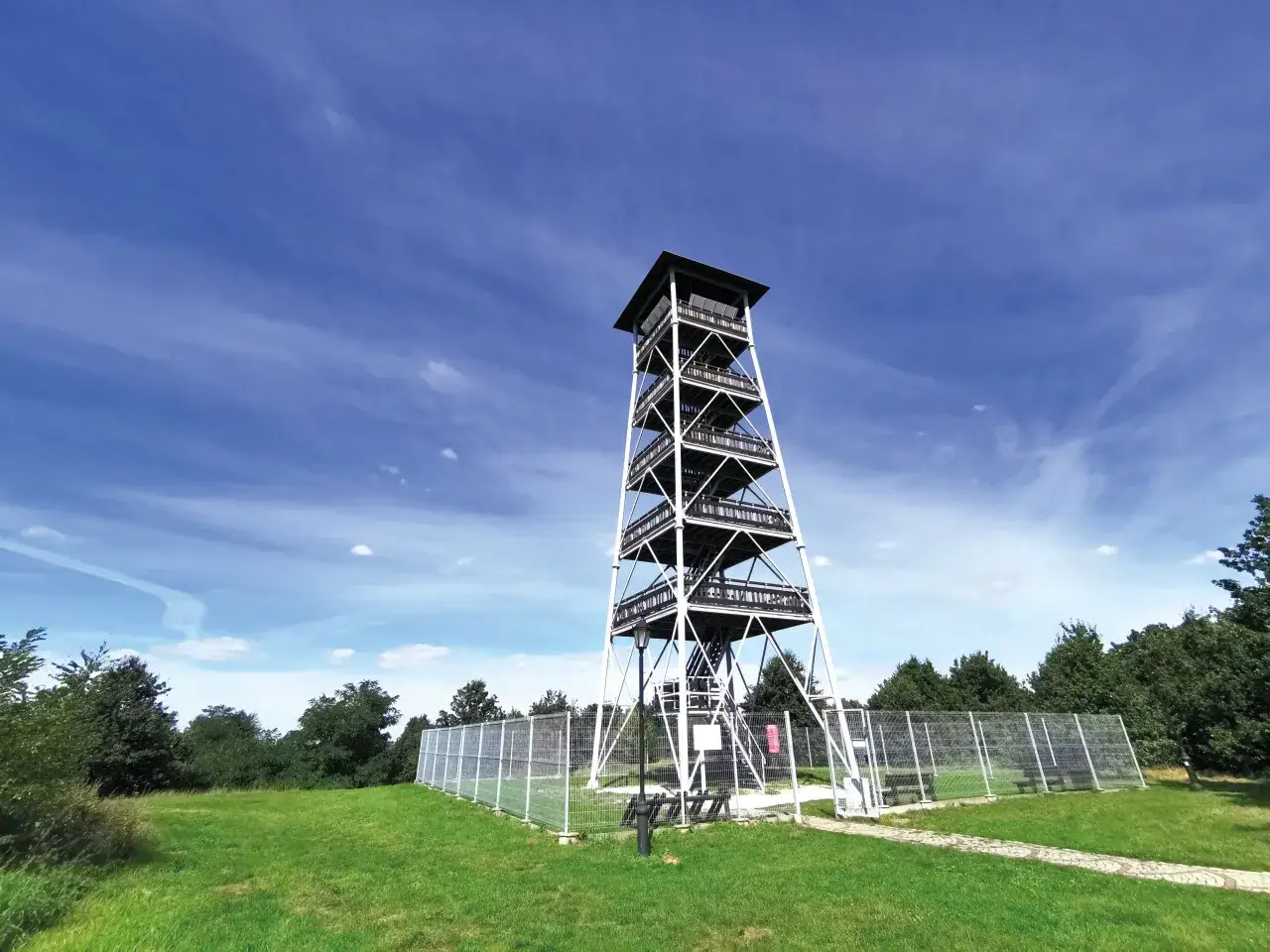 Wieża widokowa Mściwojów wznosi się na tle błękitnego nieba z delikatnymi chmurami. Otoczona zieloną trawą i ogrodzeniem, zaprasza do podziwiania panoramy.