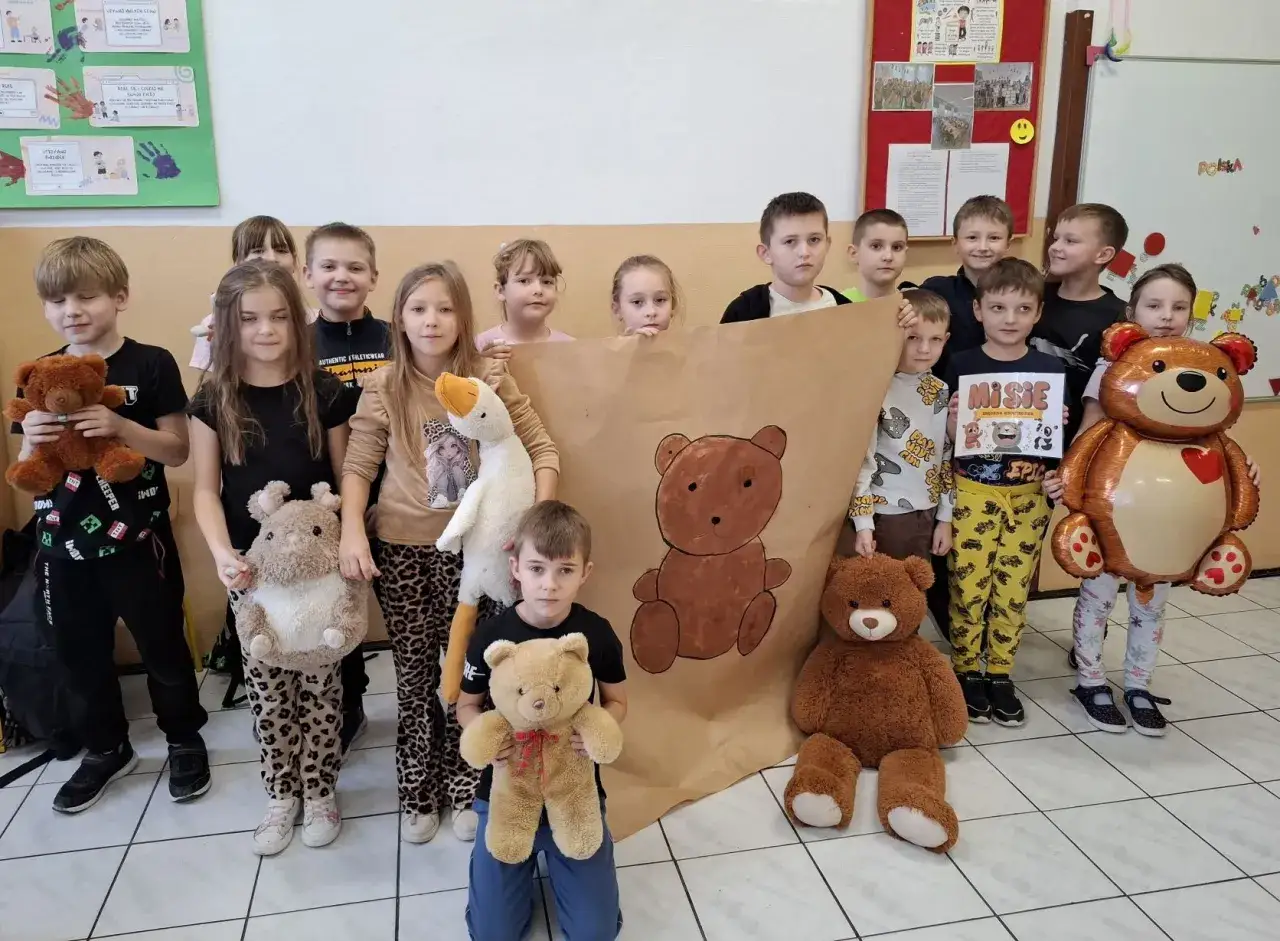Dzieci świętują dzień misia, trzymając pluszaki i wielkiego, dmuchanego misia. Na tle rysunek misia i plakat z napisem 