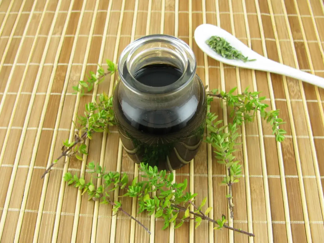 Szklany słoiczek z ciemnym płynem, otoczony gałązkami tymianku. Obok na łyżeczce suszone liście tymianku.