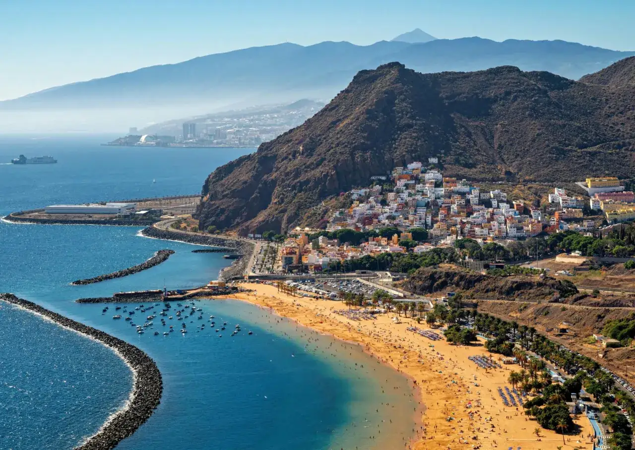 Widok na plażę i miasteczko, gdzie widać, że czy w Hiszpanii jest morze, to zdecydowanie tak! Błękitna woda, łodzie i góry w tle.
