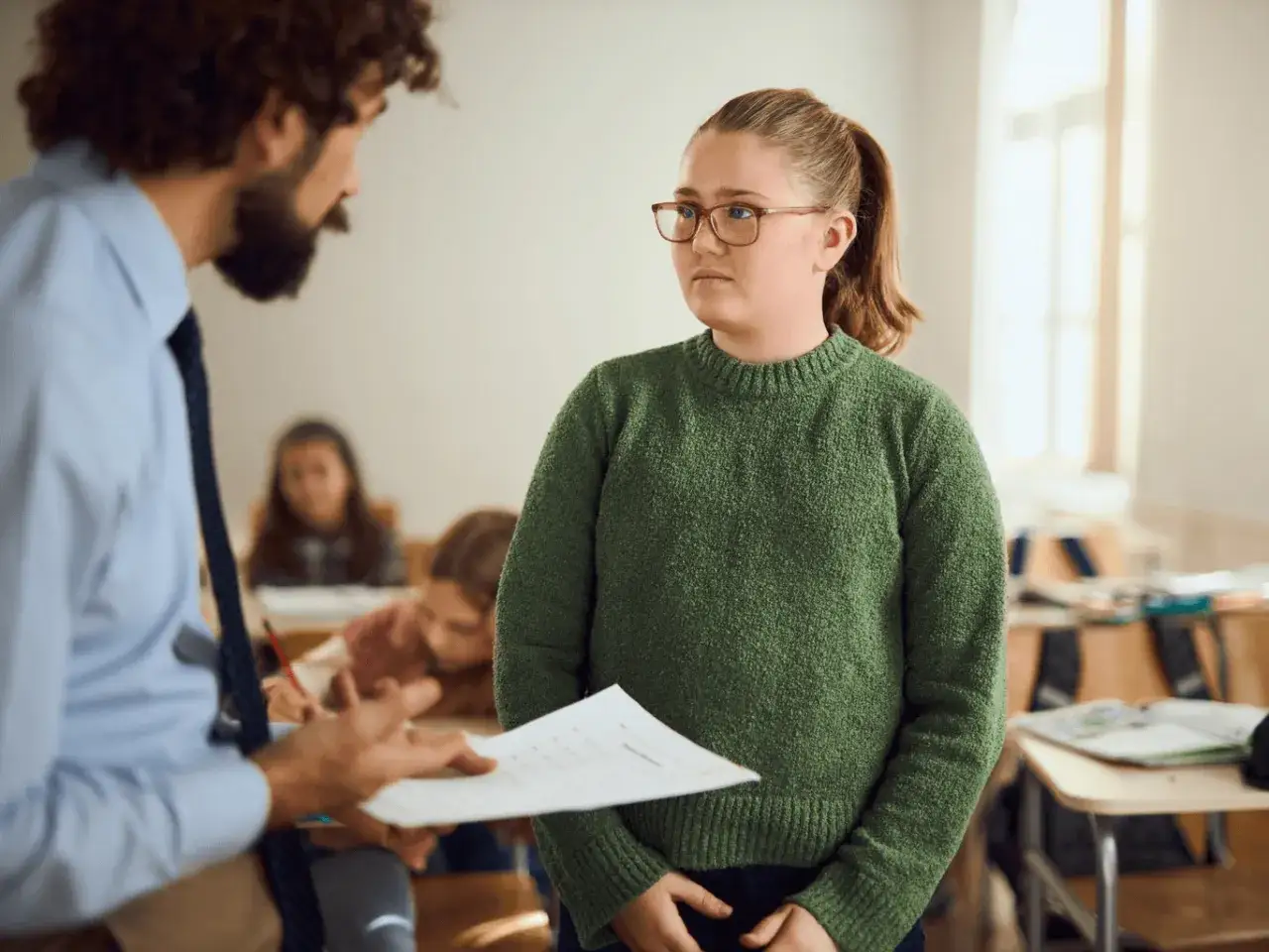 Nauczyciel rozmawia z uczennicą, która wygląda na smutną. W tle inni uczniowie, sugerując problem przemocy rówieśniczej w szkole.