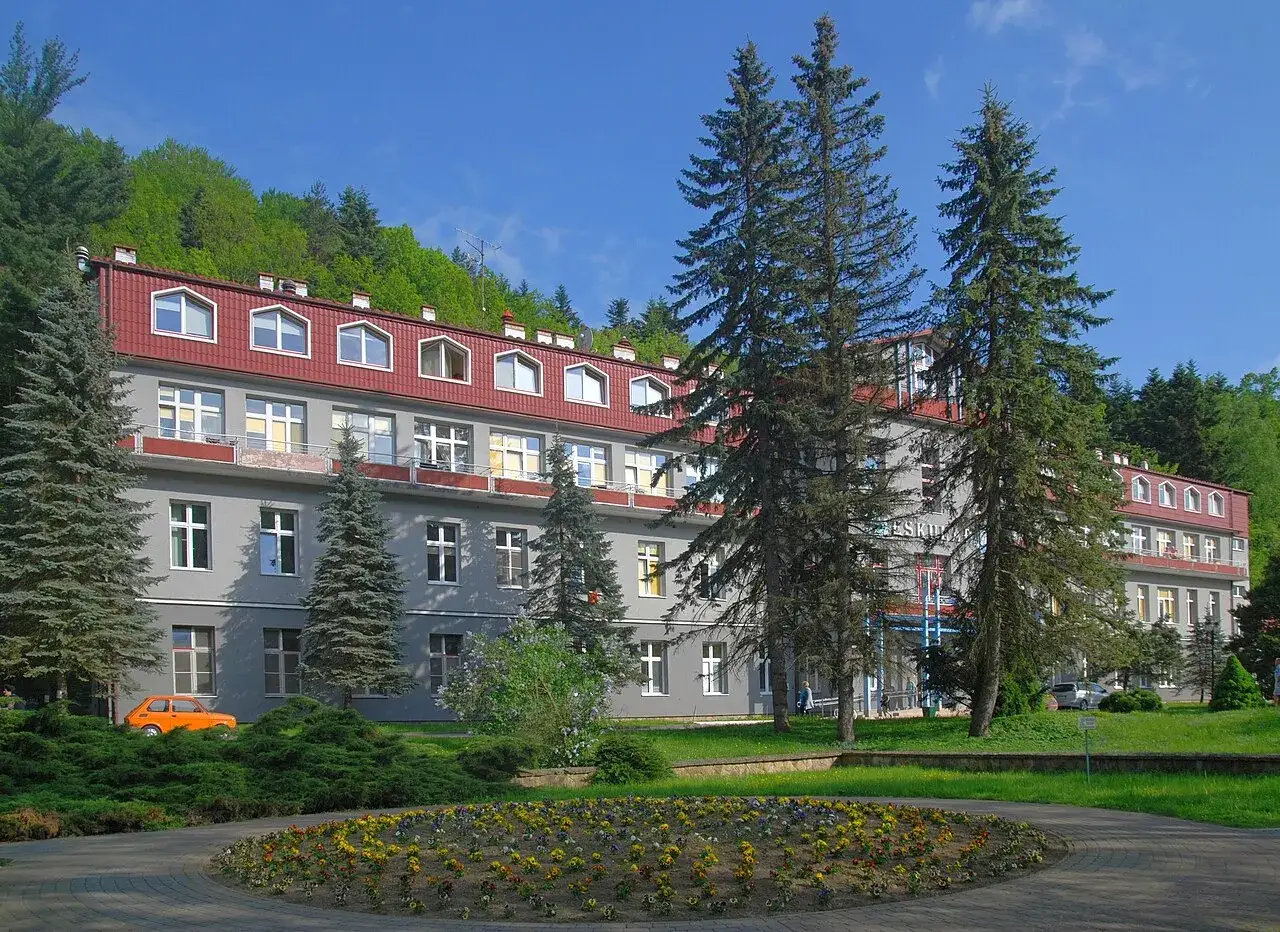 Sanatoria w Rymanowie Zdroju otoczone zielenią. Widać budynek z czerwonym dachem, drzewa i kwitnące rabaty.