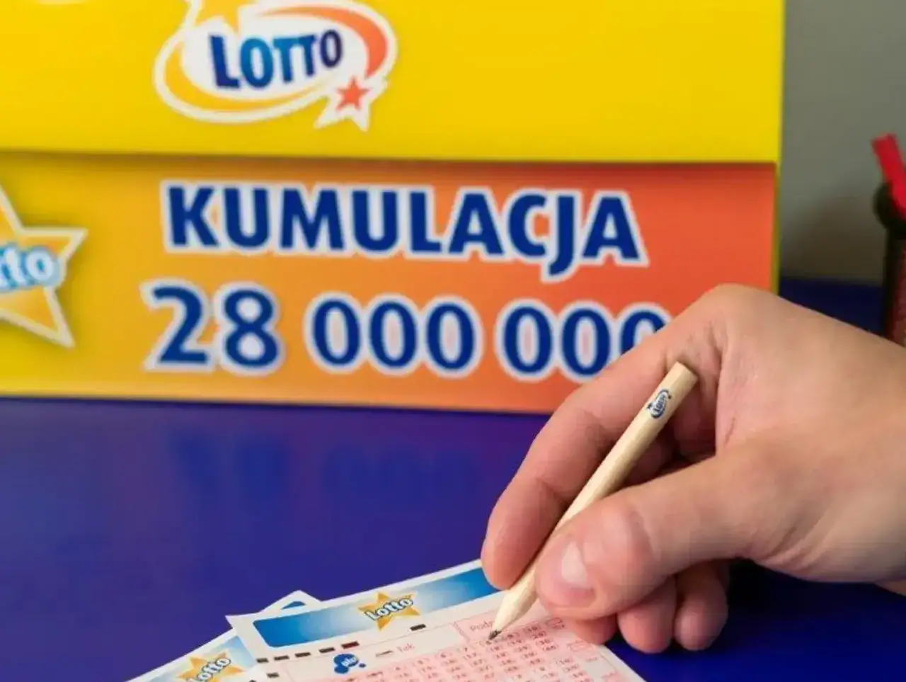 Ręka z ołówkiem nad kuponem Lotto. Za nią baner z napisem 