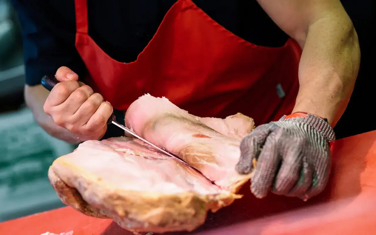 Rzeźnik w czerwonym fartuchu i rękawicy z siatki metalowej precyzyjnie kroi mięso, pokazując, jak pozbyć się błon i ścięgien z mięsa proste triki.