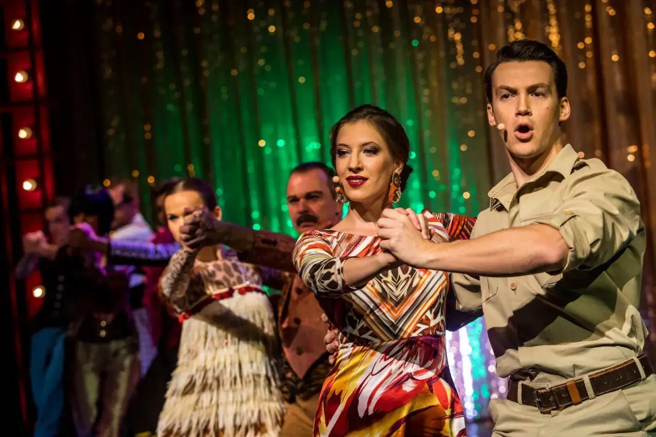 Aktor w stroju żołnierza królowej Madagaskaru tańczy z kobietą w barwnej sukni na tle zielonej kurtyny.