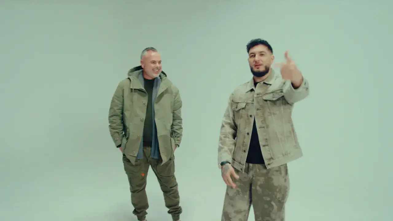 Dos hombres posan. Uno viste una chaqueta verde y un pantalón cargo. El otro, con un pantalón de camuflaje, señala a la cámara.