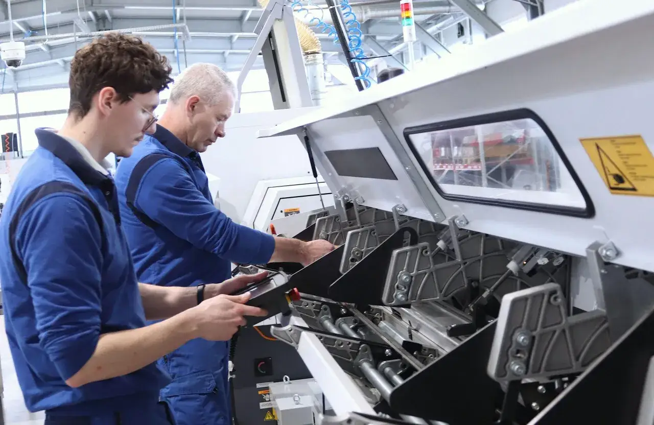 Dw&oacute;ch mężczyzn w niebieskich kombinezonach pracuje przy maszynie. Jeden z nich trzyma tablet, drugi pochyla się nad mechanizmem, prawdopodobnie zajmując się programowaniem maszyn CNC.
