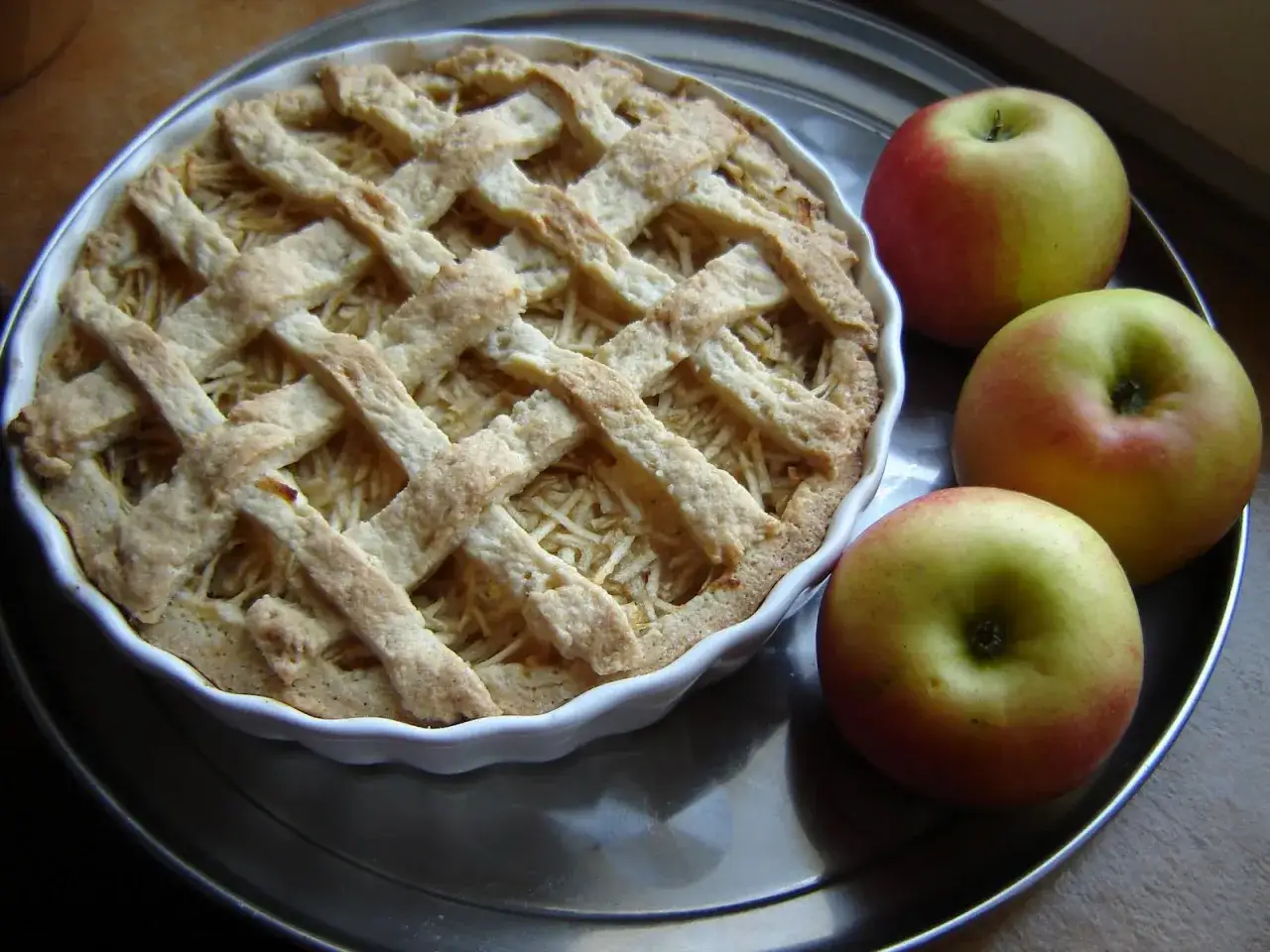 Jabłecznik z kratką z ciasta kruchego na oleju. Obok trzy rumiane jabłka.