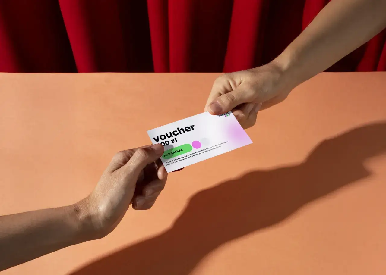 Dwie dłonie przekazują sobie voucher o wartości 100 zł. To idealny prezent na spektakl, który można wykorzystać w teatrze.