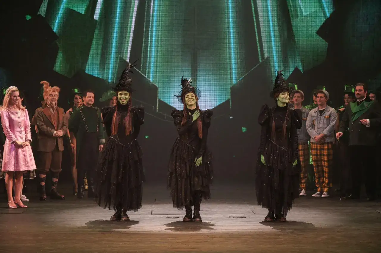 Trzy czarownice w czarnych sukniach i kapeluszach na scenie, w tle zielone światła. Spektakl 