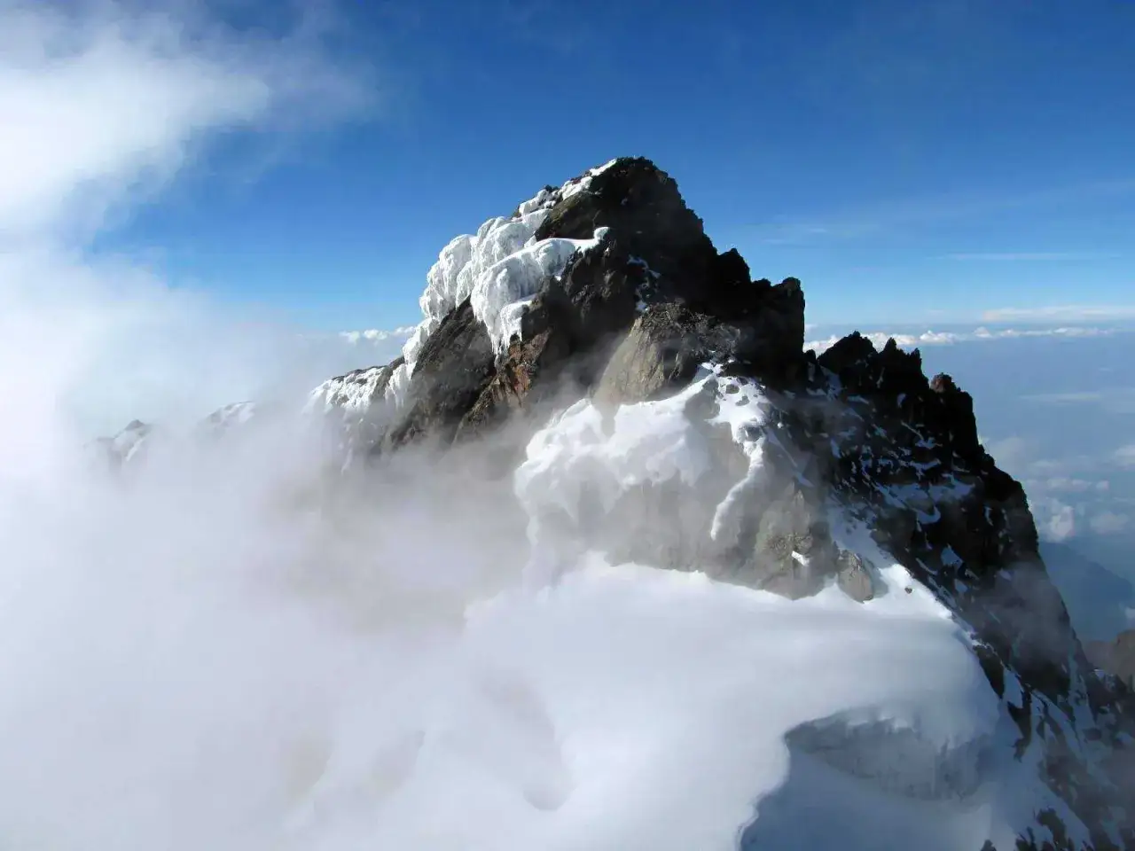 Lodowy szczyt g&oacute;ry Stanleya, otoczony chmurami, lśni w słońcu.
