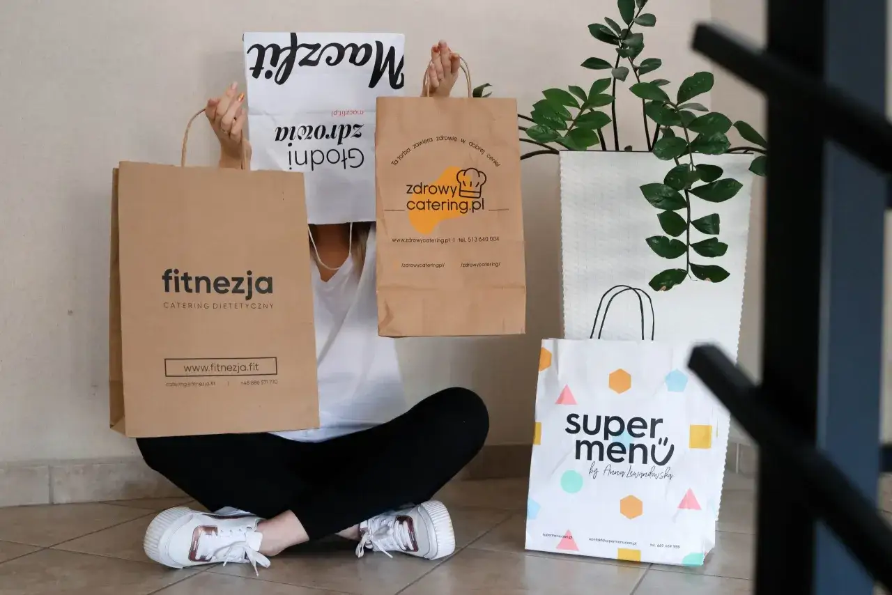 Osoba z torbami cateringowymi: Maczfit, Fitnezja, Zdrowy Catering, Super Menu. Szukasz najlepszych firm oferujących catering dla biur w dużych miastach?