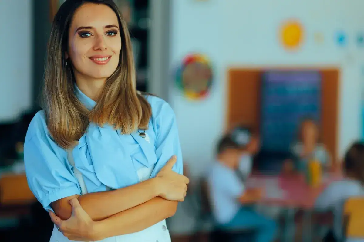 Uśmiechnięta nauczycielka w jasnoniebieskiej koszuli, z rękami skrzyżowanymi na piersi. W tle widać dzieci przy stolikach, co sugeruje, że czy nauczyciel geografii może uczyć przyrody.