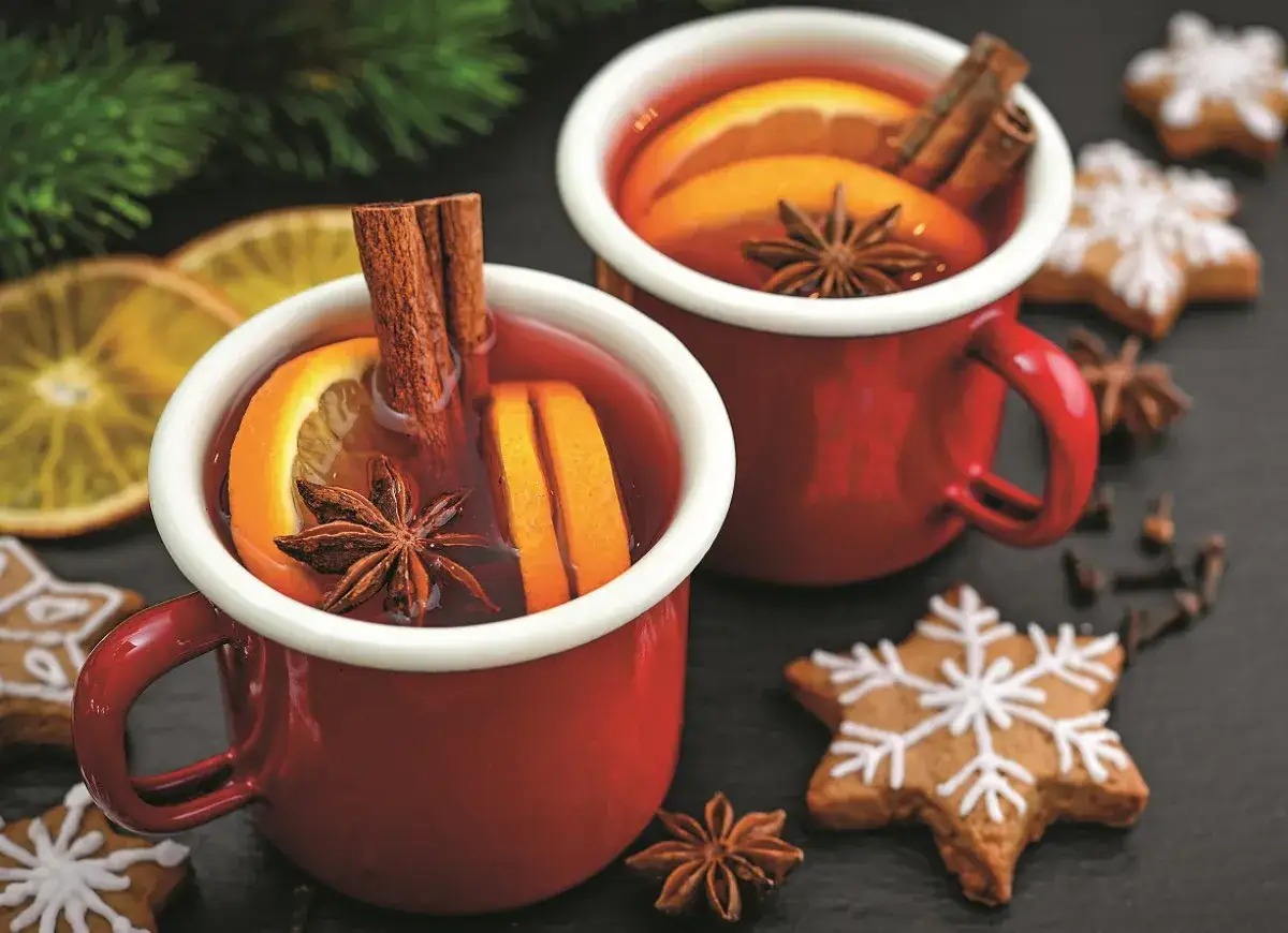 Dwa czerwone kubki z grzanym winem, ozdobione pomarańczą, cynamonem i anyżem. Obok świąteczne pierniczki i gałązka choinki. Idealna przyprawa do grzanego wina.