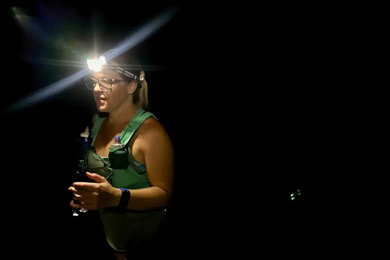 Kobieta w okularach i kamizelce z bidonami, oświetlona mocną czoł&oacute;wką do biegania, przygotowuje się do nocnego treningu.