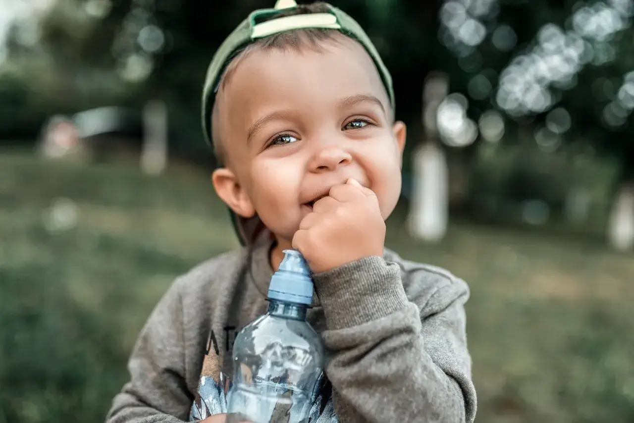 Uśmiechnięty chłopiec w czapce z daszkiem trzyma butelkę z wodą i wkłada palec do ust. To może być odpowiedź na pytanie, dlaczego dziecko gryzie przedmioty.