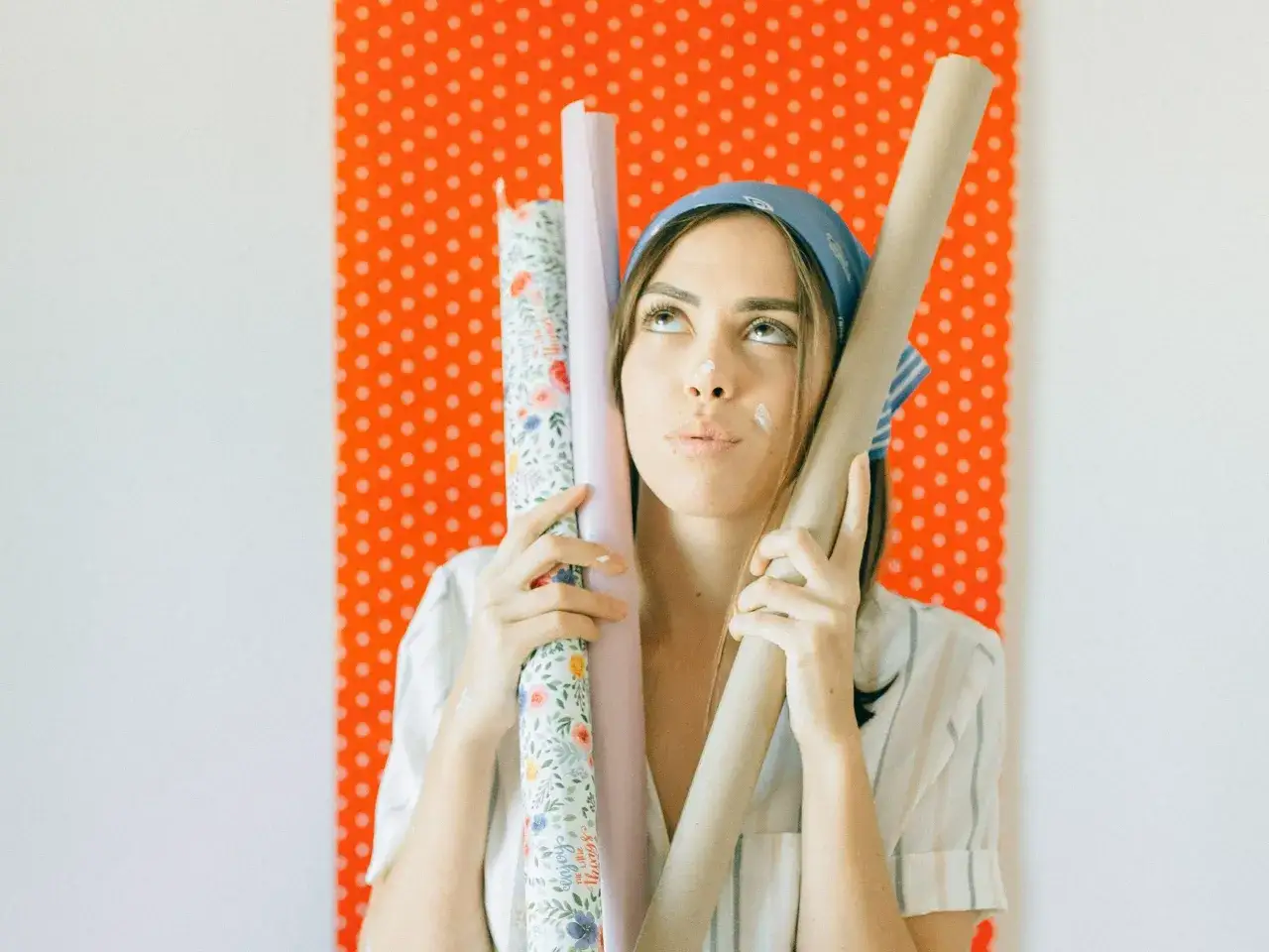 Kobieta z wałkami tapety zastanawia się, jak położyć tapetę. W tle pomarańczowa ściana w kropki.