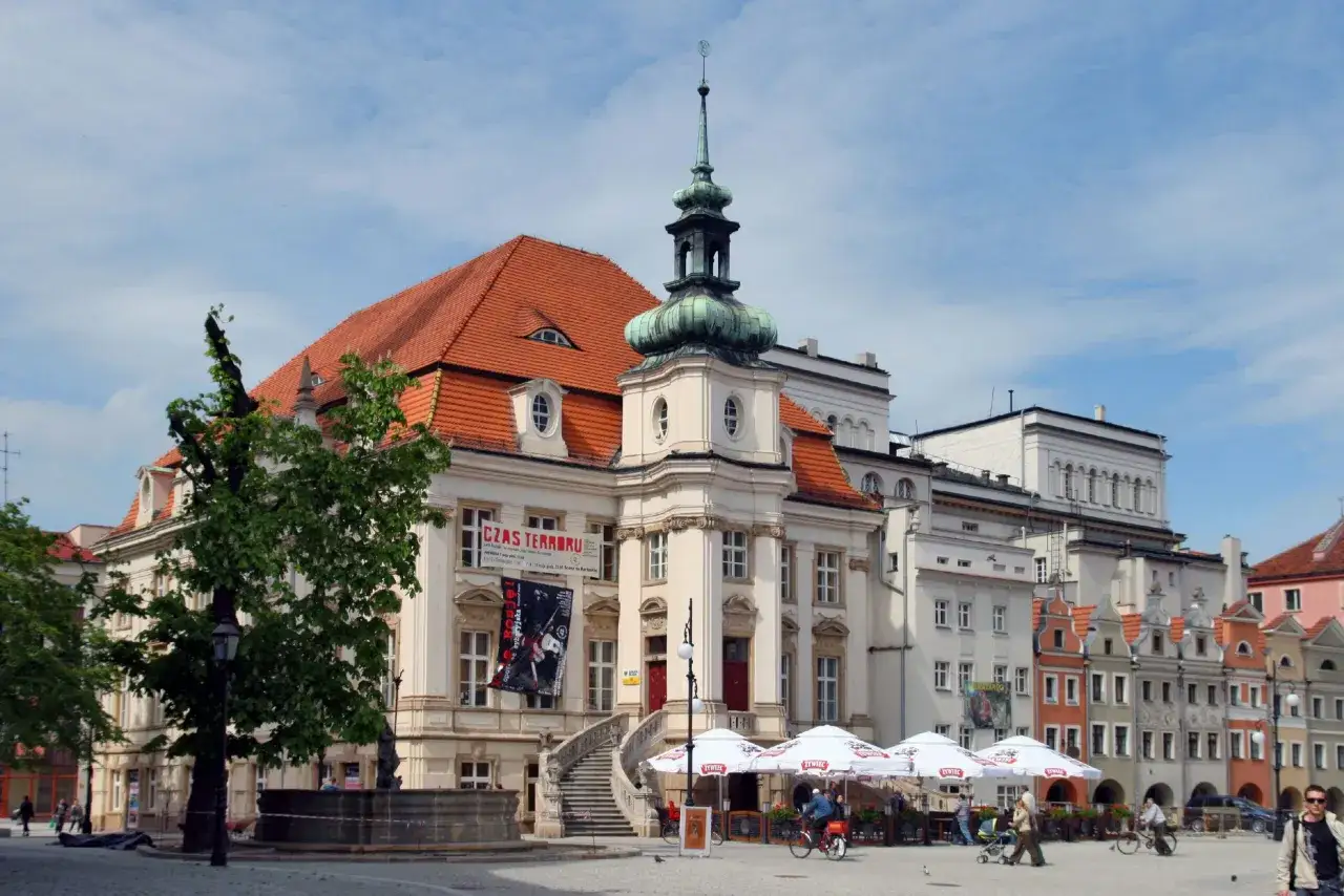 Rynek w Legnicy z ratuszem i kolorowymi kamieniczkami. Widać kawiarniane parasole i ludzi spacerujących po placu. To jedne z wielu legnickich atrakcji.