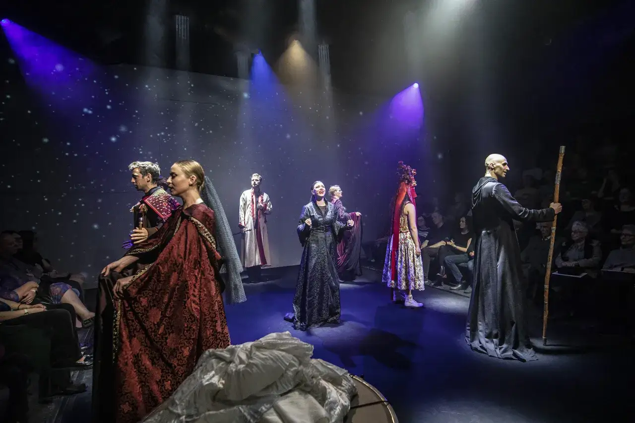Maria Tyszkiewicz w teatralnej scenografii, w otoczeniu aktorów w barwnych strojach, na tle gwieździstego nieba.