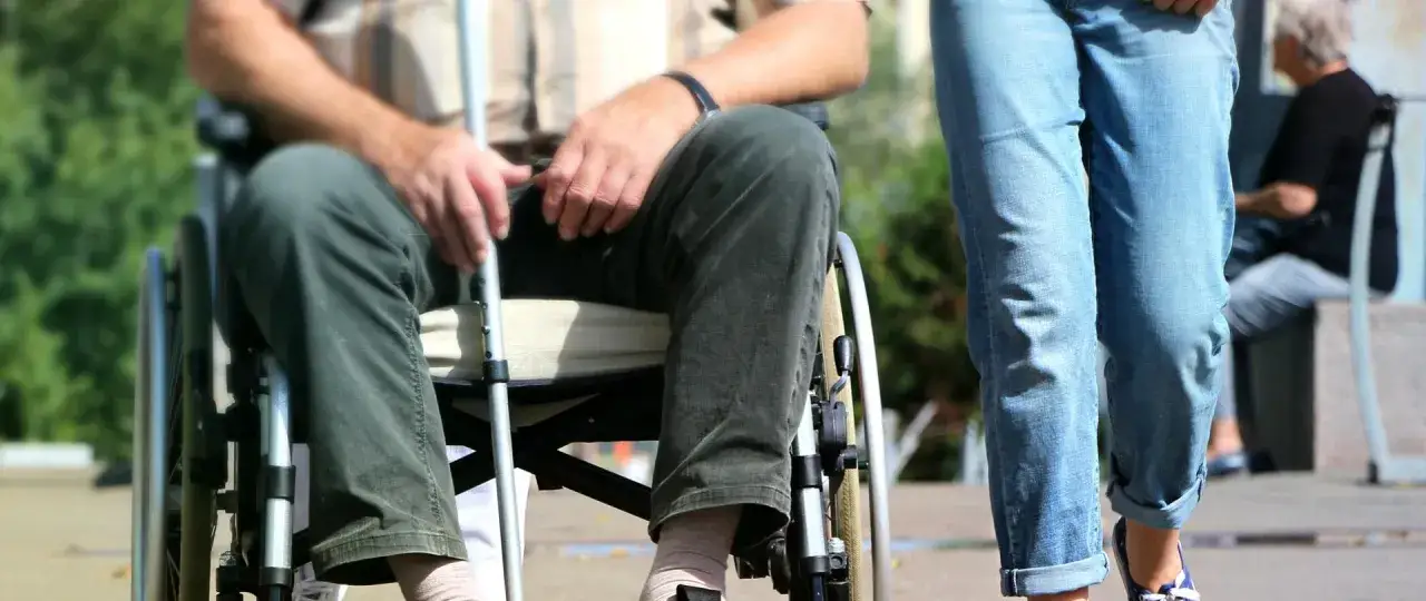 Osoba na wózku inwalidzkim z laską, obok niej idzie druga osoba. W tle siedzi starsza osoba. Dostępne są szczególne uprawnienia dla osób ze znacznym stopniem niepełnosprawności.