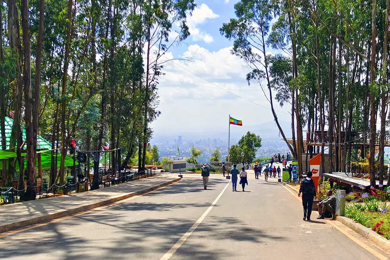 Widok na Addis Abeba z zielonego wzg&oacute;rza, gdzie powiewa etiopski sztandar. Ludzie spacerują alejką wśr&oacute;d drzew.