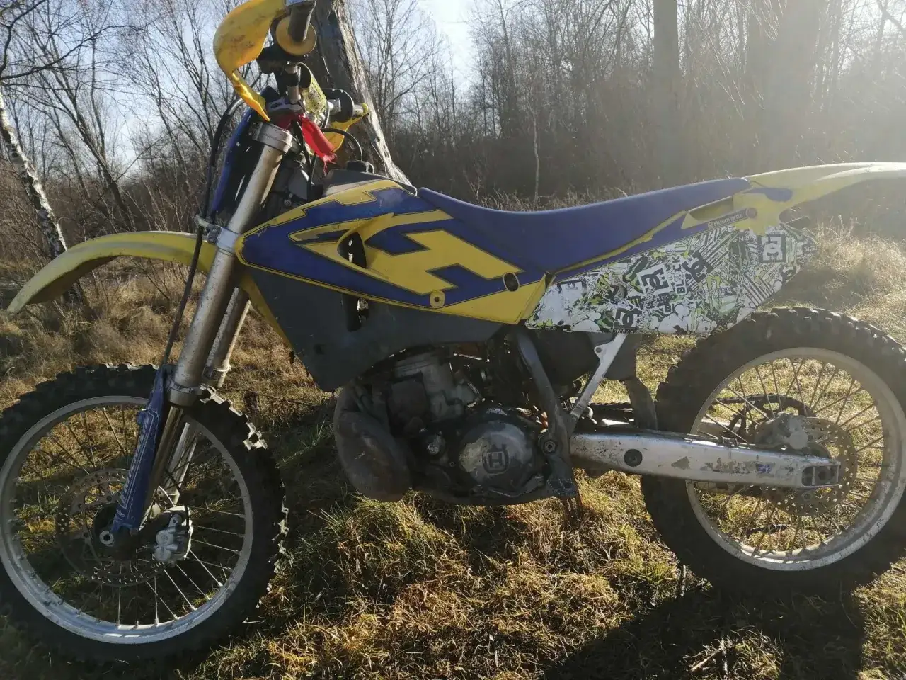 Husqvarna CR 250 gotowy do jazdy po leśnych ścieżkach. Ż&oacute;łto-niebieska maszyna z naklejkami na błotniku.