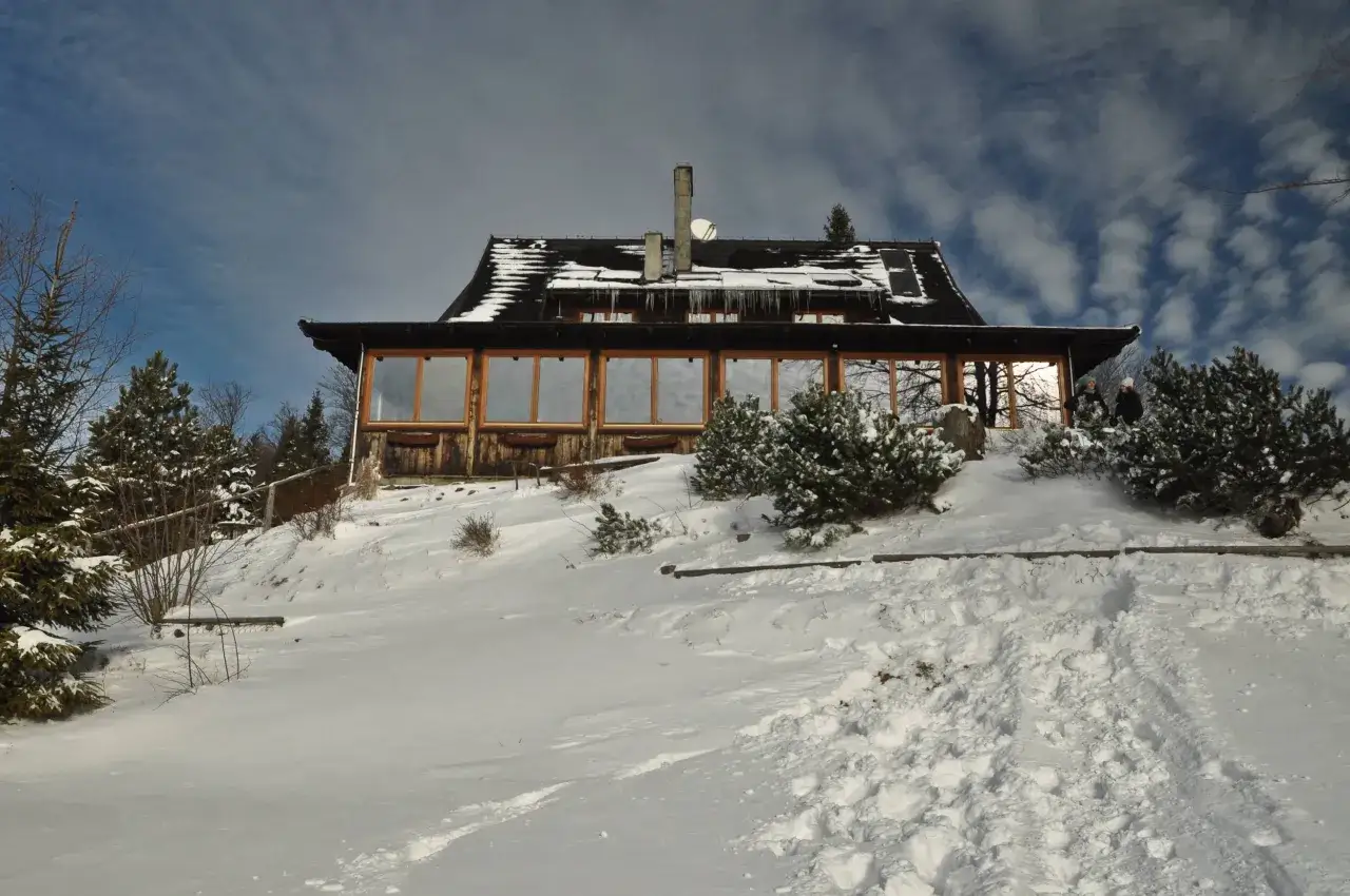 Zimowy krajobraz wok&oacute;ł bac&oacute;wki PTTK nad Wierchomlą. Śnieg pokrywa wzg&oacute;rze, a na dachu schroniska wiszą sople.