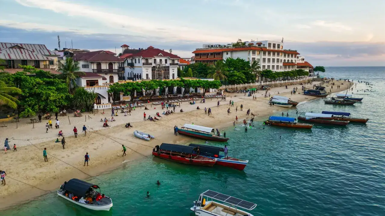 Plaża na Zanzibarze, gdzie turyści odpoczywają, a łodzie czekają na rejsy. W tle hotel i budynki, a w wodzie widać ludzi. Waluta na Zanzibarze to szyling tanzański.