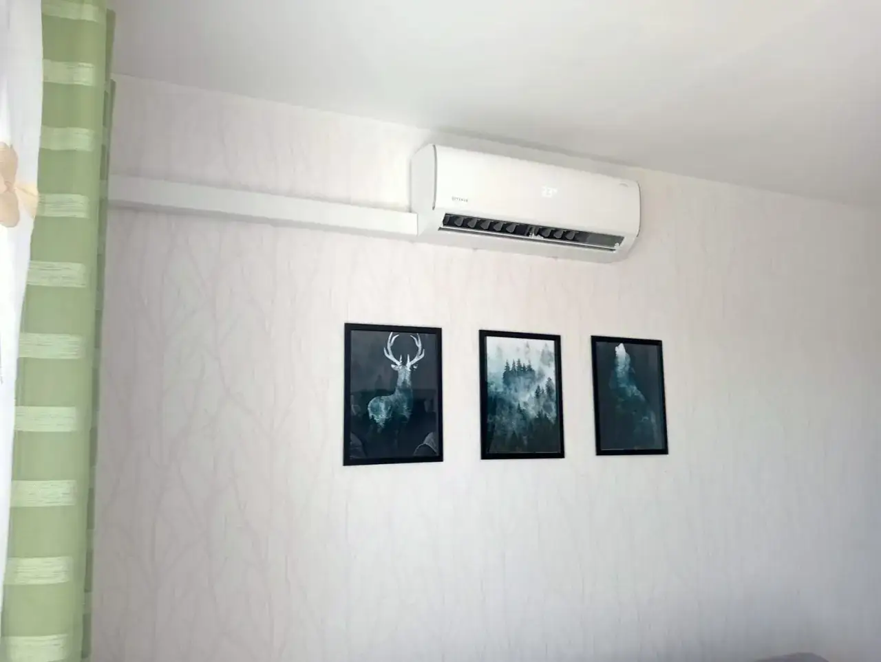 Biała klimatyzacja w mieszkaniu, zamontowana nad trzema obrazami z motywami leśnymi.
