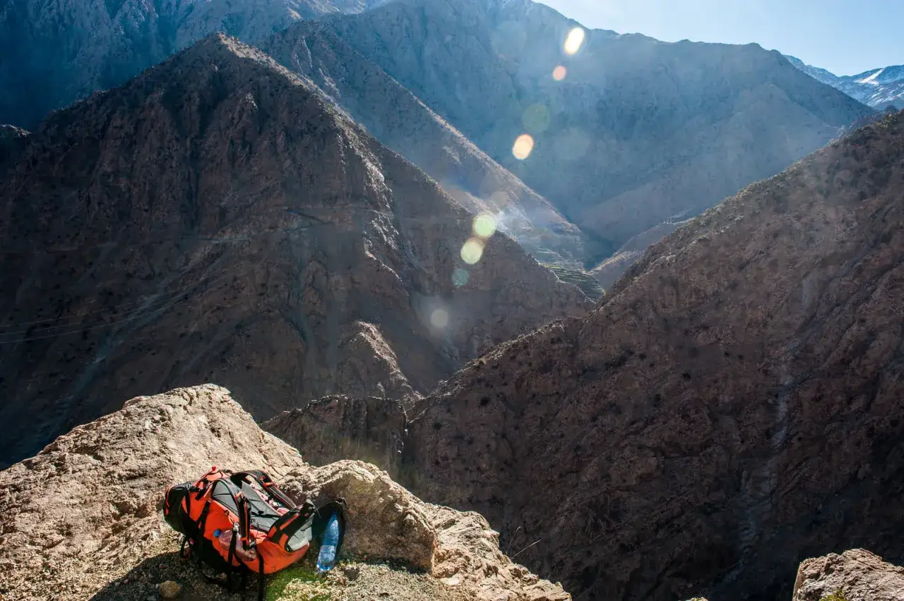Pomarańczowy plecak i butelka wody na skale w g&oacute;rach Atlas w Maroku. Widok na skaliste zbocza i doliny.