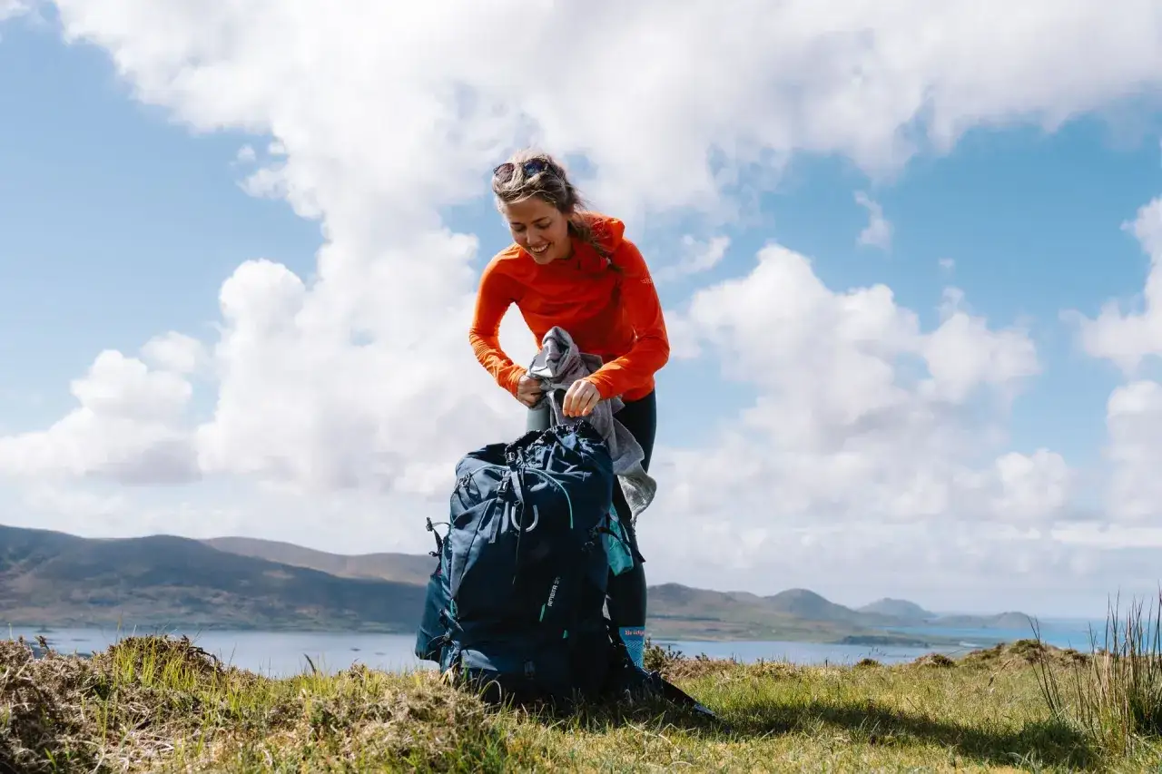 Kobieta w pomarańczowej bluzie pakuje plecak na tle g&oacute;r i jeziora. Właśnie dowiedziała się, jak prać plecak, by był gotowy na kolejną wyprawę.