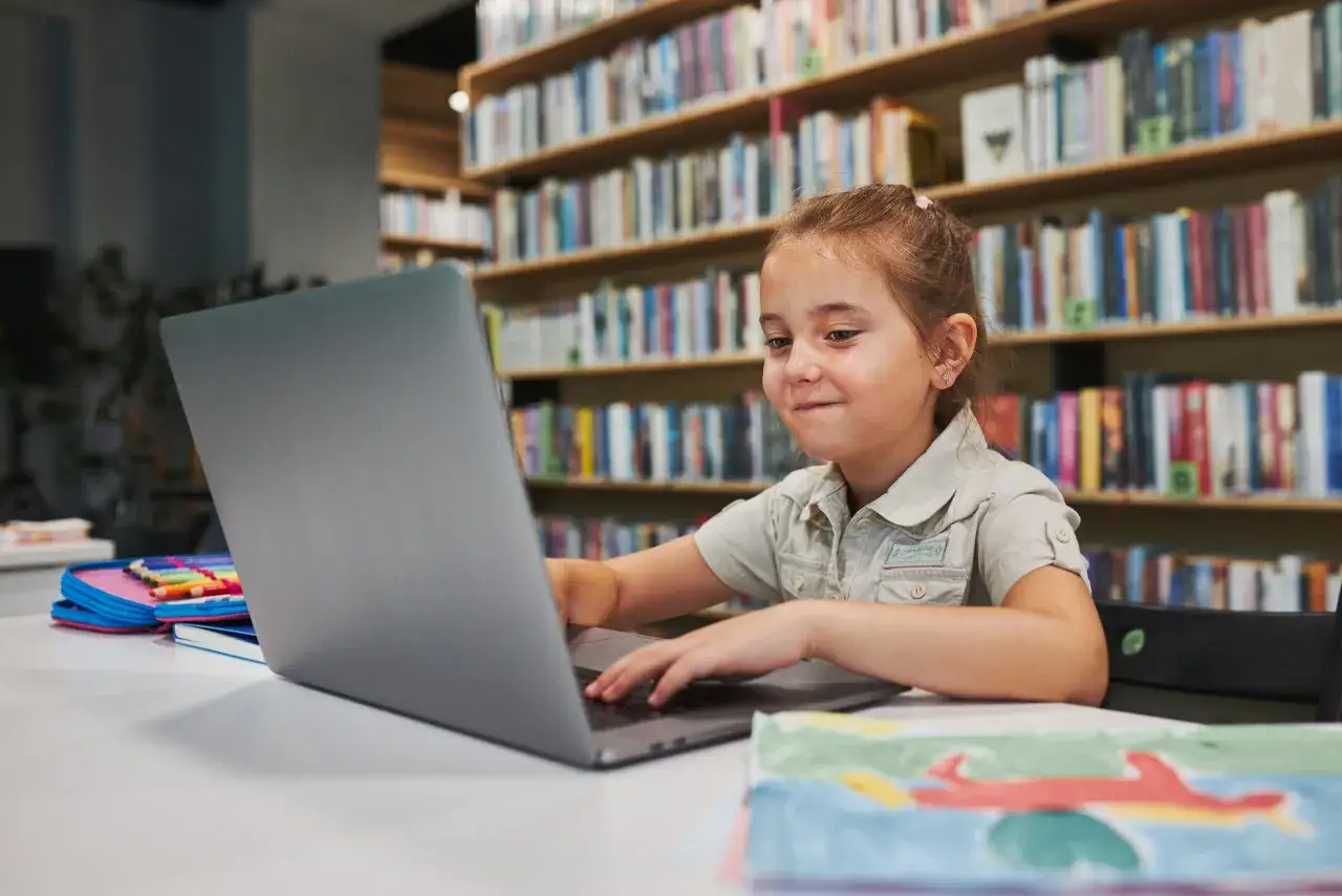 Dziewczynka w bibliotece uczy się, jakie przedmioty są w 4 klasie. Na stole ma laptopa, pi&oacute;rnik z kredkami i zeszyt.