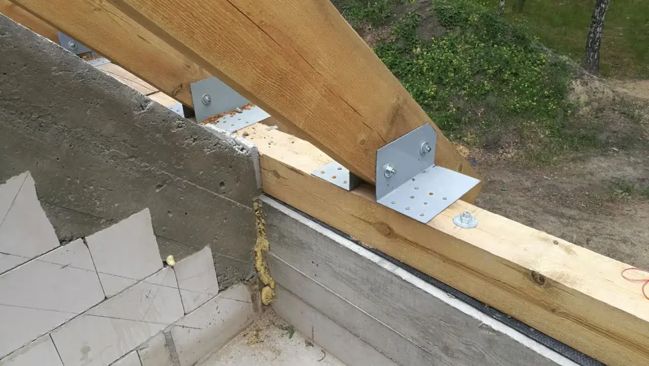 Drewniana konstrukcja dachu opiera się na betonowym murze. Widoczne są metalowe kątowniki mocujące krokwie do murłaty, która stanowi podstawę dachu.