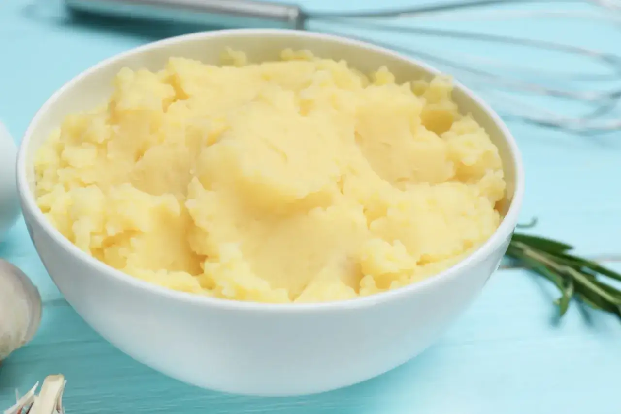 Puszyste puree ziemniaczane w białej misce, idealne do zagęszczenia. Obok czosnek i rozmaryn.