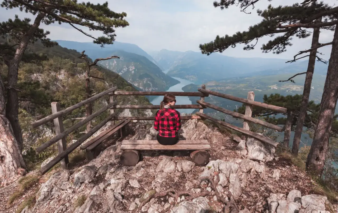 Kobieta w kratę siedzi na ławce na szczycie góry w Serbii, podziwiając malowniczy widok rzeki wijącej się w dolinie.