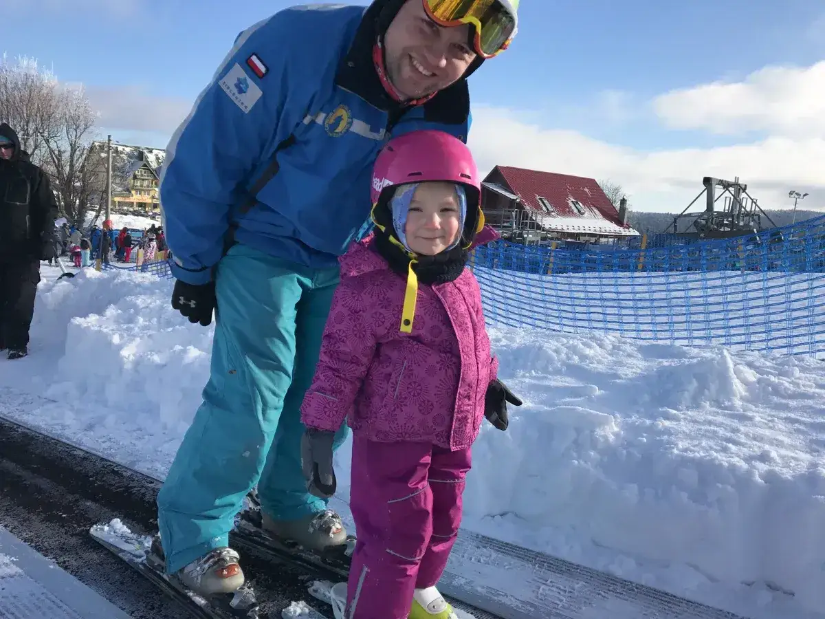 Uśmiechnięty instruktor narciarstwa pomaga dziecku na taśmie w Zieleńcu. Dowiedz się, ile kosztuje wypożyczenie sprzętu narciarskiego w Zieleńcu.