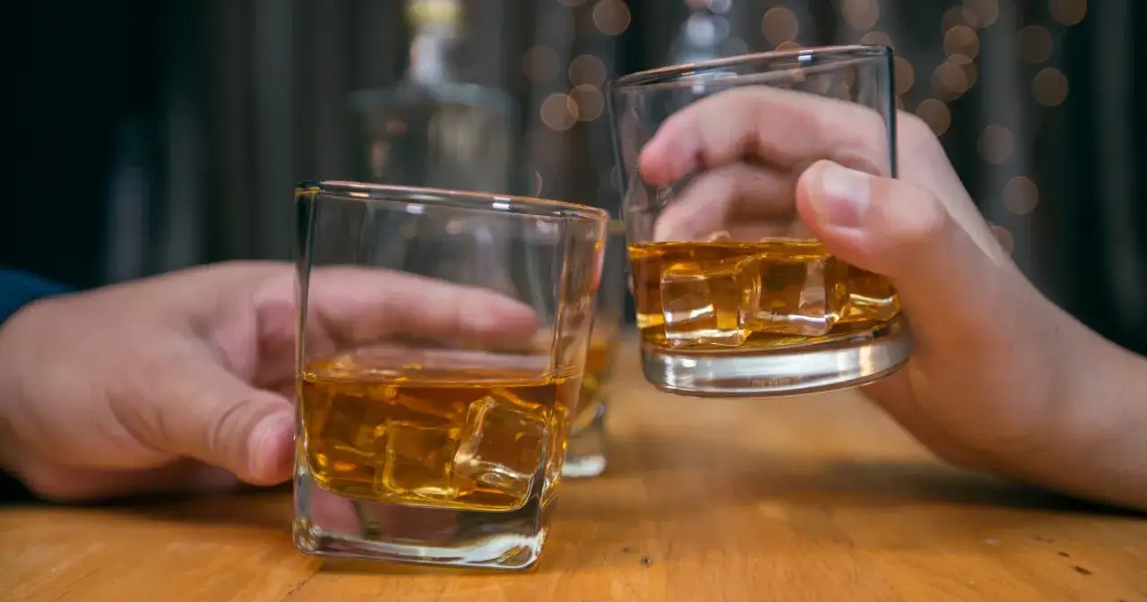 Dwie ręce unoszą szklanki z whisky i lodem. Czy whisky jest zdrowe? To pytanie towarzyszy tej chwili relaksu.