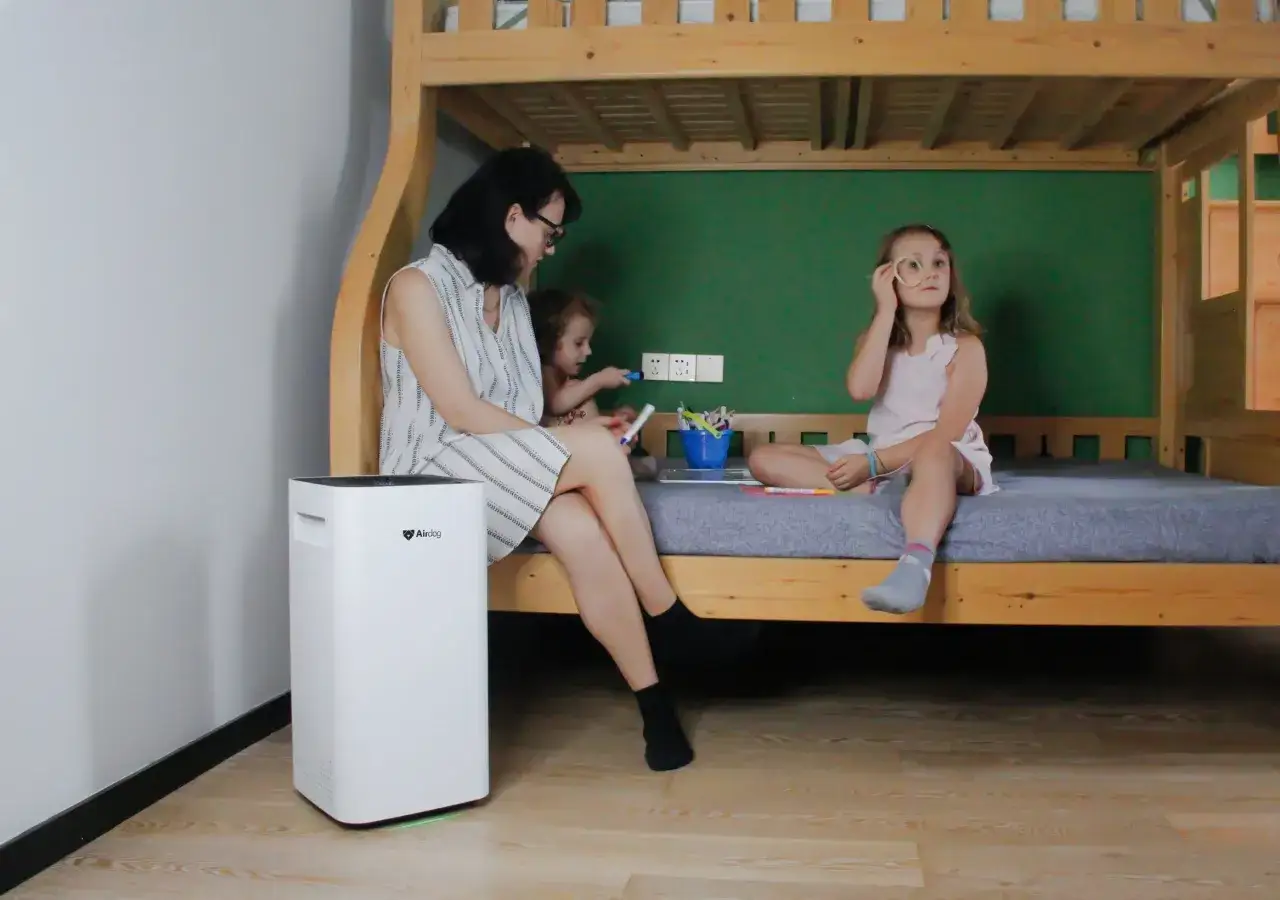 Kobieta i dzieci siedzą na ł&oacute;żku piętrowym obok białego oczyszczacza powietrza. Czy to jego wady?