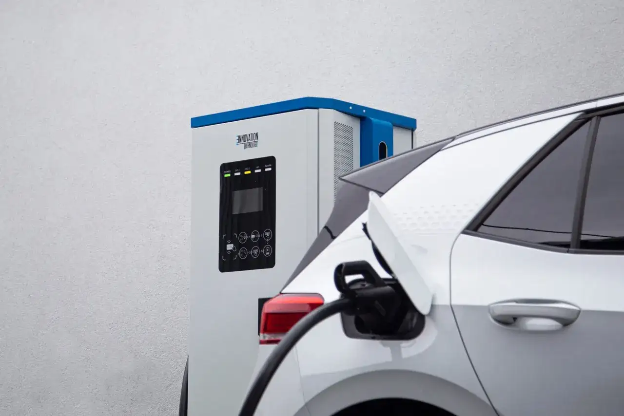 Ładowanie samochodu elektrycznego. Złącze ładowania podłączone do białego auta, obok stacja ładowania.