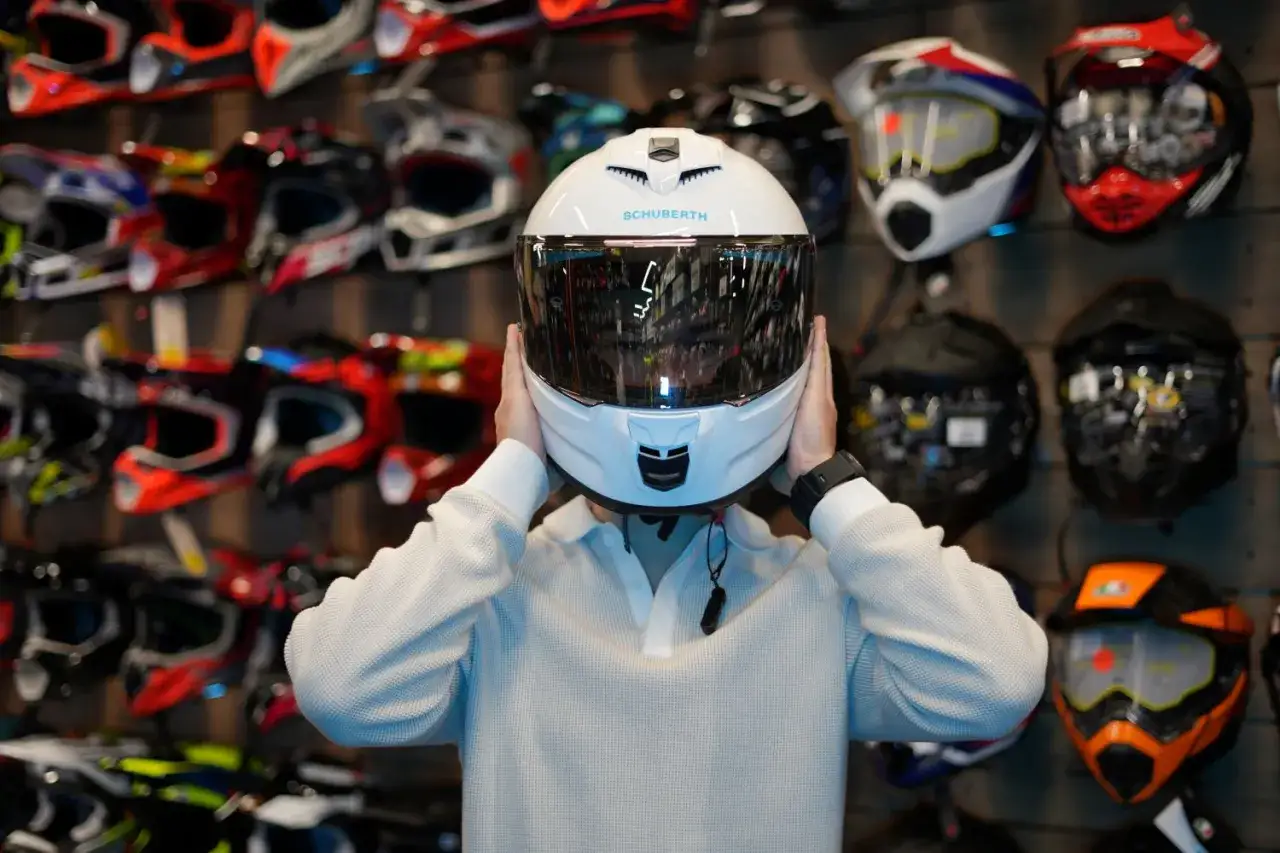 Osoba przymierza biały kask Schuberth. W tle widać różne rodzaje kasków motocyklowych, od sportowych po turystyczne.