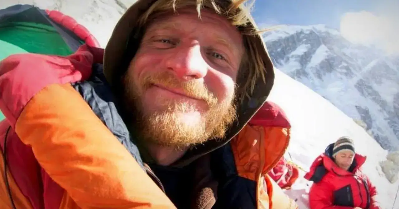 Uśmiechnięty alpinista z zarostem, w kolorowej kurtce, na tle ośnieżonych g&oacute;r. Widać też drugą osobę w czerwonej kurtce. To mogą być objawy choroby wysokościowej.