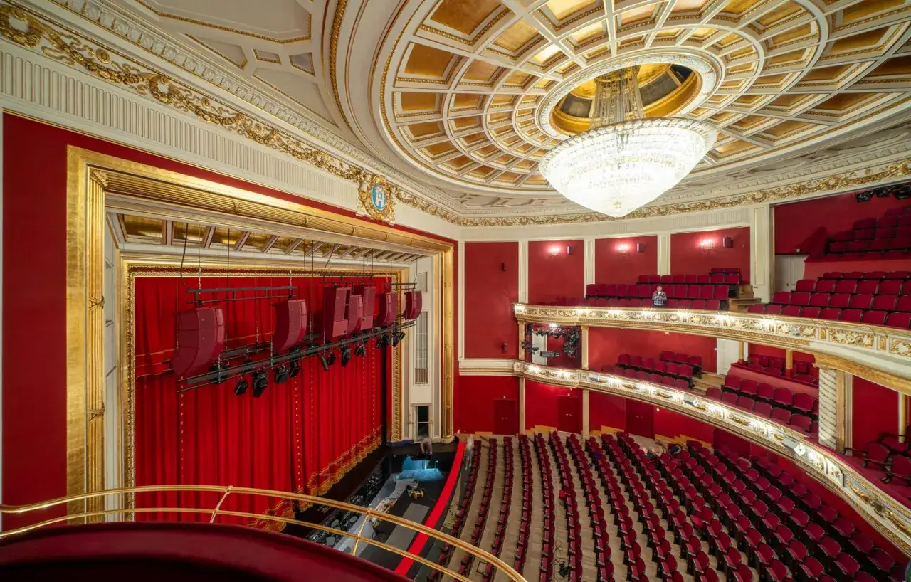 Wielki teatr, plan widowni z czerwonymi fotelami i złotymi zdobieniami. Złoty żyrandol i kurtyna zapraszają na spektakl.