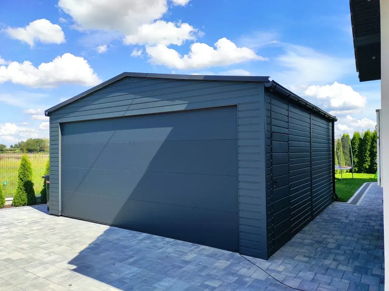 Nowy, ciemnoszary garaż z bramą segmentową stoi na podjeździe z kostki brukowej. Jest to garaż 1,5 m od granicy działki.