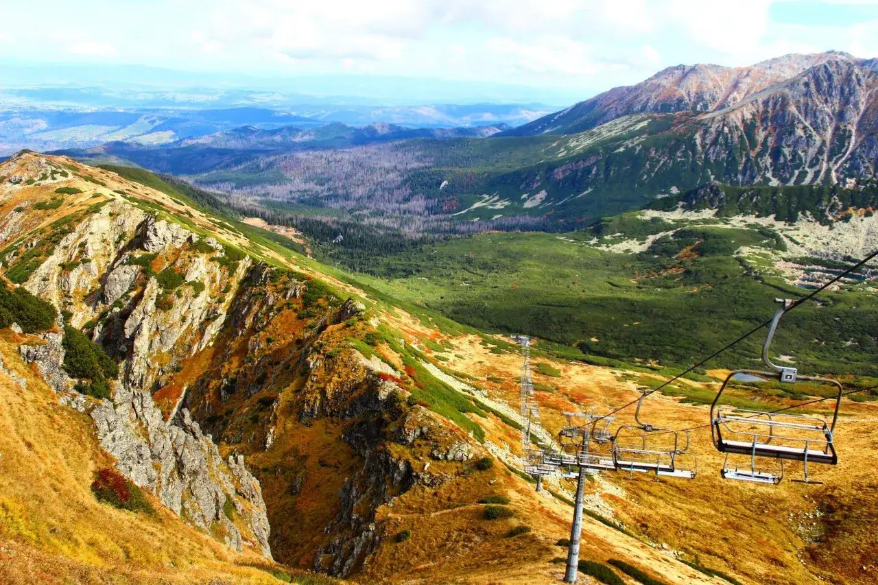 Jesień na szlaku na Kasprowy Wierch. Kolejka krzesełkowa wjeżdża na zbocze pokryte złotą trawą i zieloną kosodrzewiną. W oddali widać pasma g&oacute;rskie.