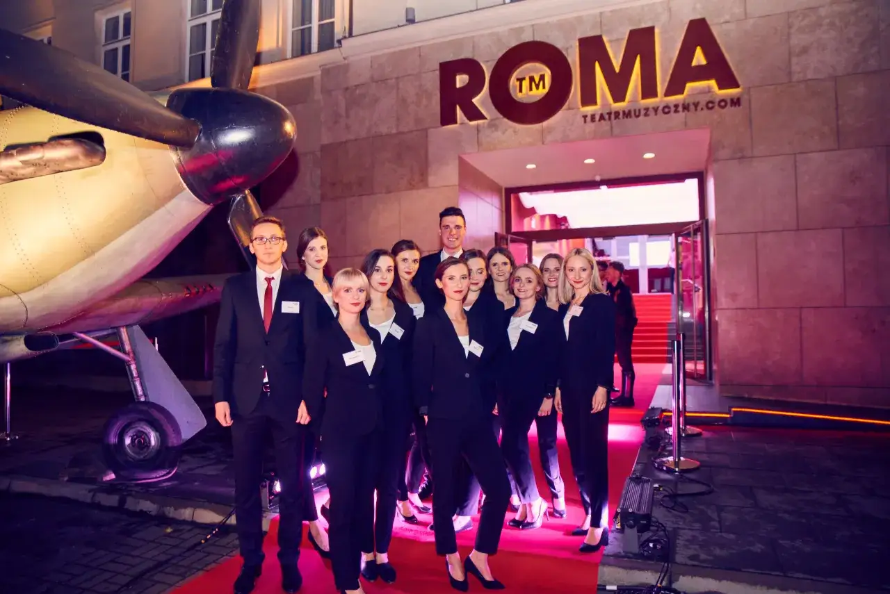 Zespół Teatru Roma w eleganckich strojach, gotowy do pracy. Obok samolot, symbol podróży i niezwykłych wrażeń.