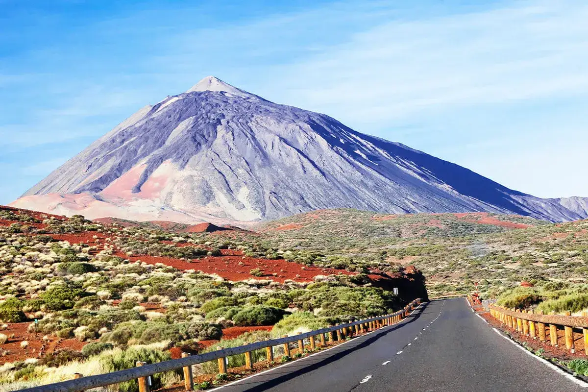 Droga prowadzi do majestatycznego wulkanu Teide. Krajobraz jest surowy, z czerwoną ziemią i niską roślinnością.