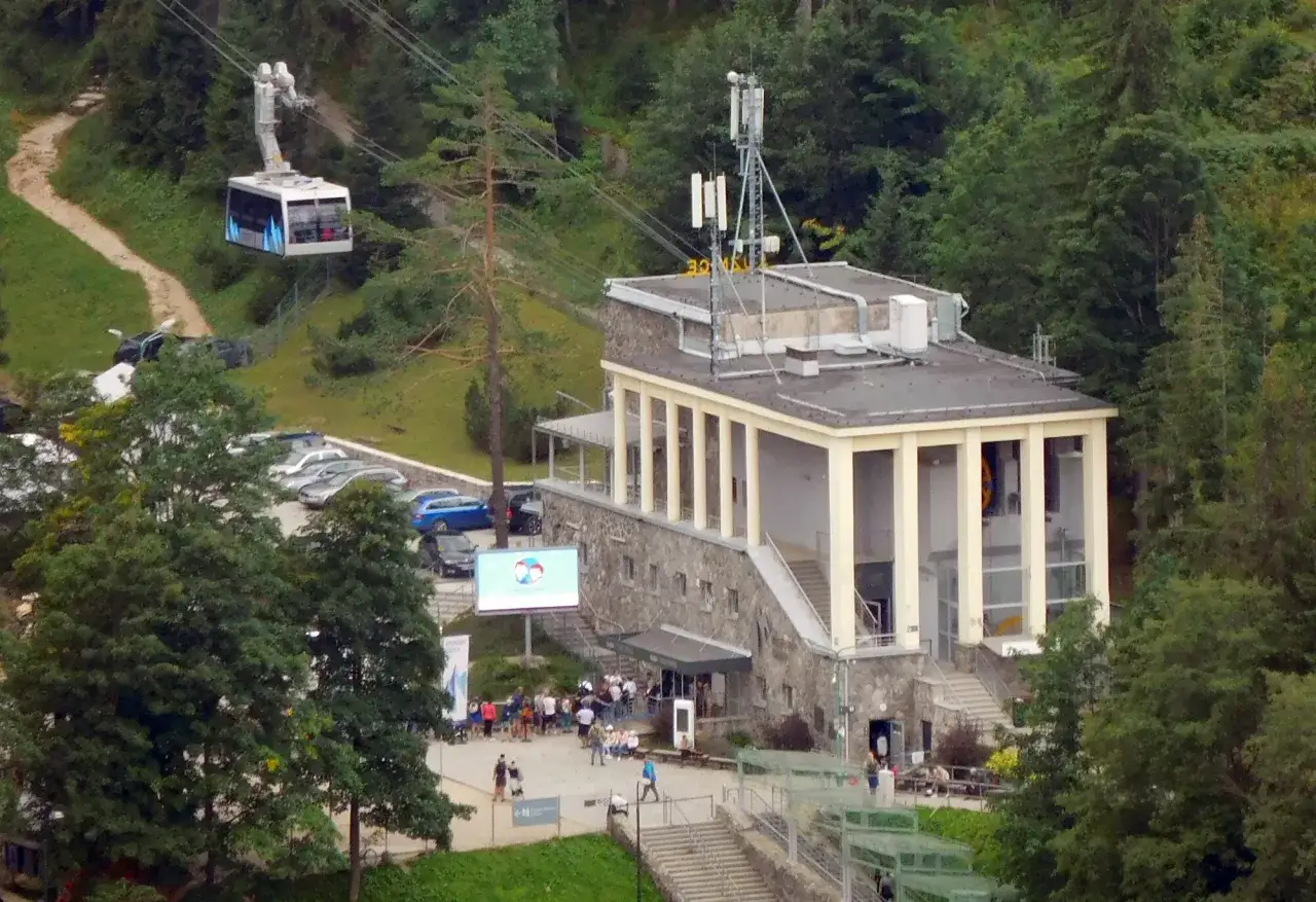 Kolejka na Kasprowy Wierch wjeżdża na g&oacute;rę. Obok budynku stacji widoczny jest parking i ludzie czekający na wejście. Sprawdź wyciąg na Kasprowy Wierch cennik.